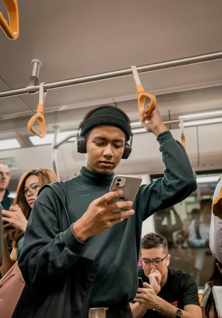 Person on a train looking at their phone with headphones over their ears; other passengers whom are also on their phones are behind them